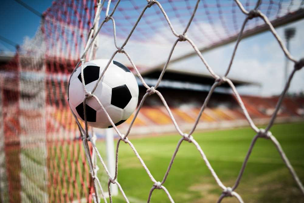 Fotoausschnitt von Arena mit Fußballfeld im Hintergrund; Nahaufnahme Fußball schießt ins Tor und prallt auf das Tornetz