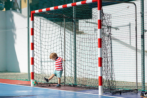 Kleiner blonder lächelnder Junge spielt Fußball. Steht im Tor und hält sich am Netz fest