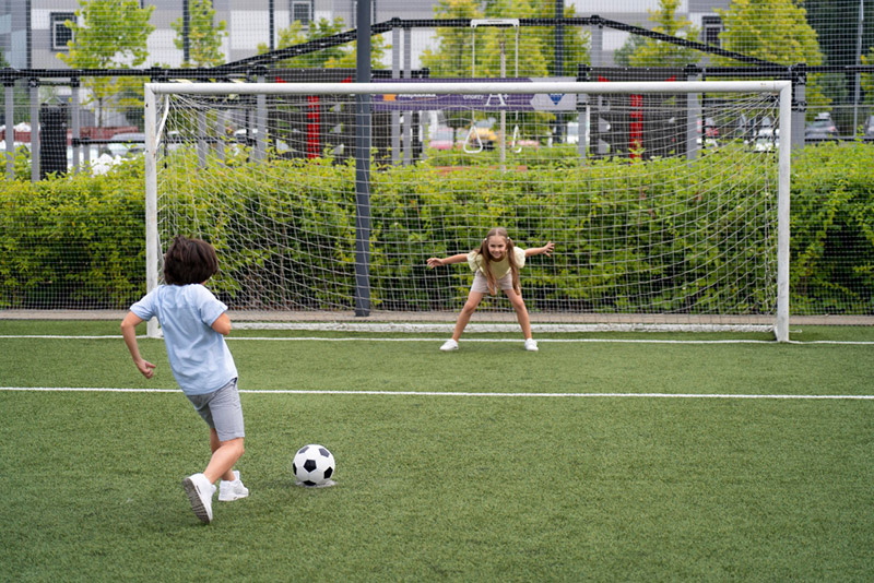 Jugendtor - Mädchen steht positioniert vor Jugendtor auf einem Sportplatz und der Junge versucht seinen Ball an ihr vorbei ins Tor zu schießen
